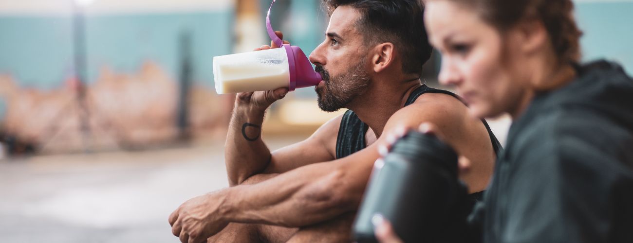 Muž a žena sedí vedle sebe po tréninku a muž pije protein z shakeru.