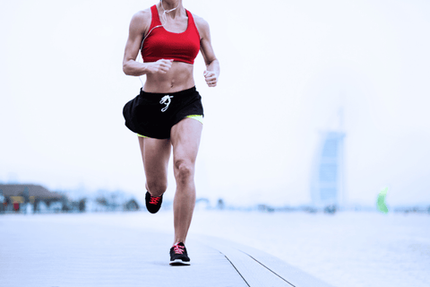 Žena běží podél mořského pobřeží v červeném sportovním topu a černých šortkách.