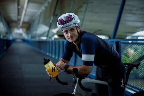 Cyklista v helmě a dresu drží láhev s iontovým nápojem hyve hydration pro během pauzy na mostě.