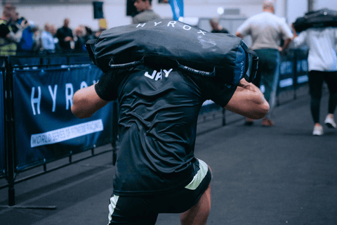 Muž v soutěži HYROX přenáší těžký pytel na ramenou.