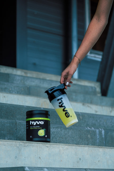 Ruka sahá po shakeru s nápojem hyve vedle dózy clear whey protein na schodech stadionu