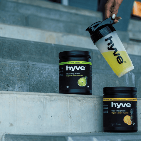 Ruka sahá po shakeru s nápojem hyve vedle dózy clear whey protein na schodech stadionu