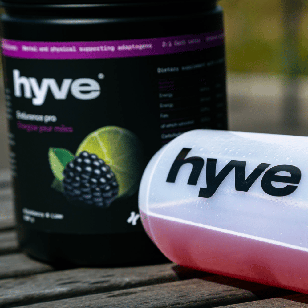 Předtréninkový nápoj pro vytrvalce hyve Endurance Pro ostružina a limetka a bidon s růžovým nápojem na venkovním dřevěném stole
