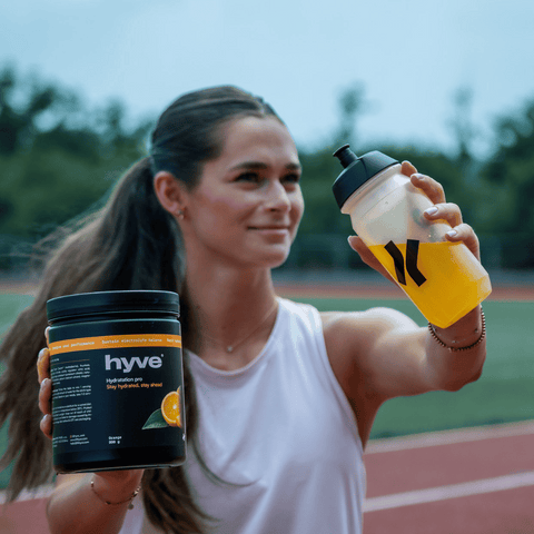 Sportovkyně drží hyve Hydration Pro s příchutí pomeranče a bidon s iontovým nápojem na atletické dráze