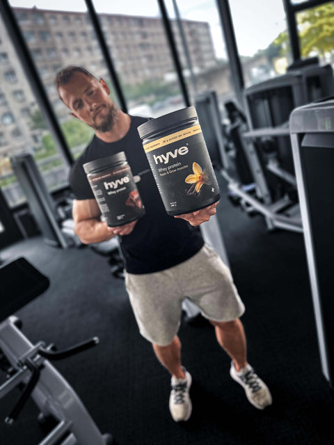 Muž ve fitku drží dvě balení proteinu hyve whey proteinu – vanilkovou a čokoládovou příchuť.