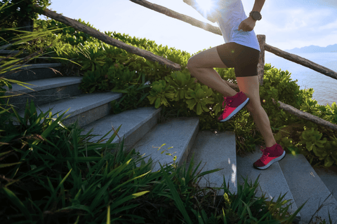 Běžkyně v růžových teniskách vybíhá schody u pobřeží obklopené zelení.